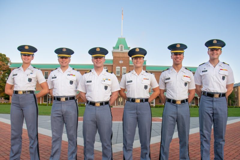 The six cadets are in their white shirt uniforms on Upper Quad. They are smiling and Lane Hall is behind them.  