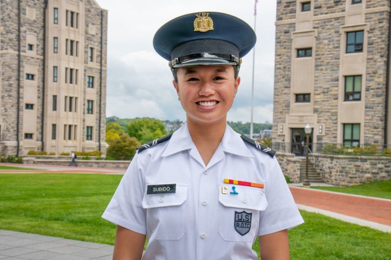 Cadet Subido stands in her uniform smiling in front of Hokie stone buildings. 
