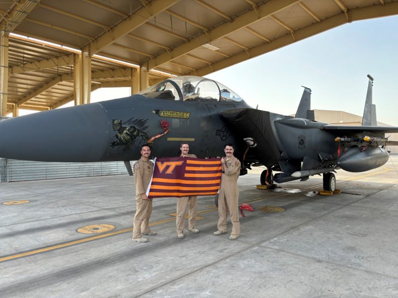 three officers stand in front of a military fighter jet while holding a striped VT flag. They are smiling and wearing flight suits. 