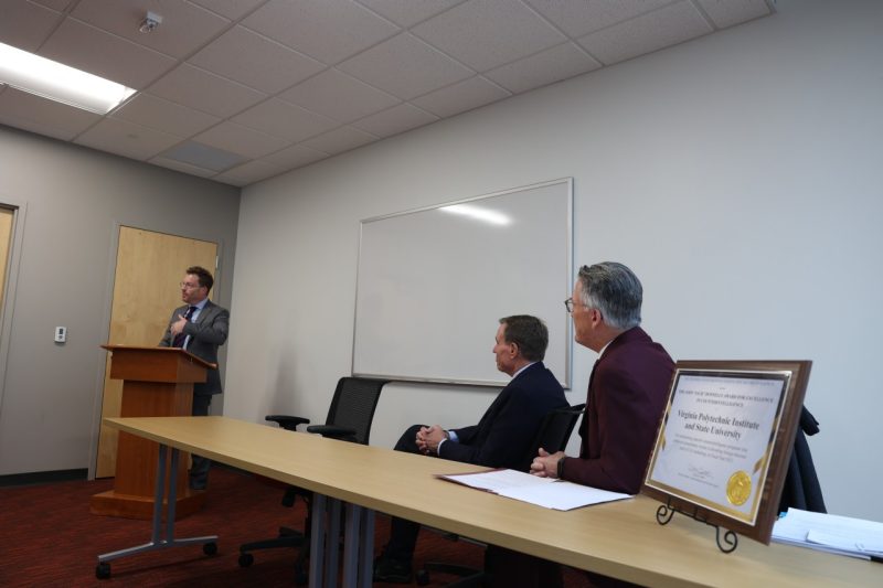 Person speaking at podium as two people look on from seats at a nearby table