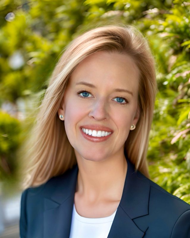 Woman with blonde hair in a blue jacket and a white shirt smiling.