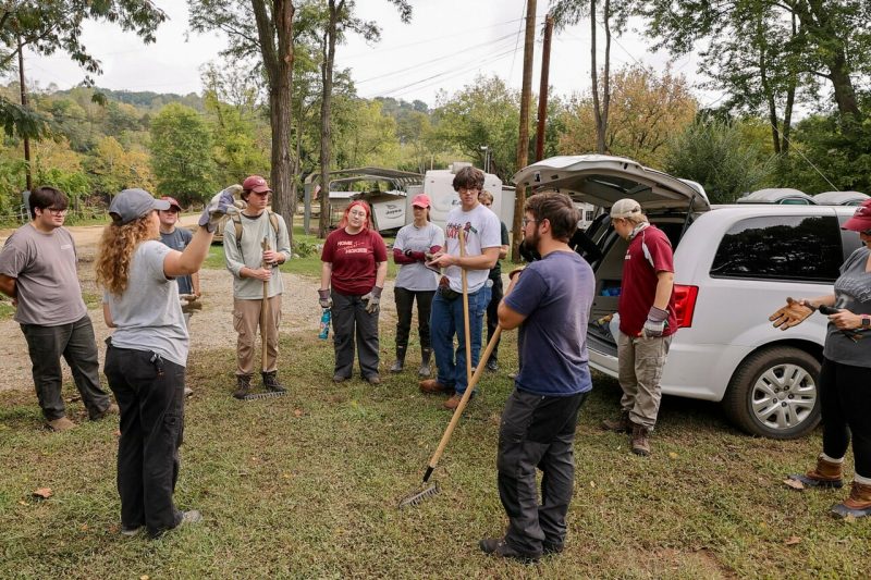 A group of VT Engage volunteers stands in a circle at a campground in Giles County, VA. Many volunteers are wearing gloves, and some are holding rakes and other handheld tools. Izzy Largen, assistant director for food access initiatives with VT Engage, gestures with one hand to something out of frame.