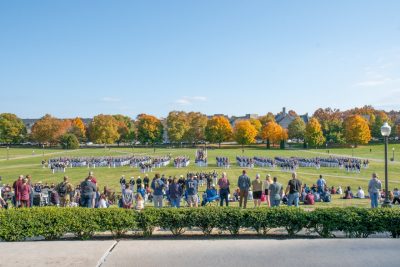 The cadet regiment stands in formation in dress uniforms on the Drillfield during a formal pass in review. Spectators line the foreground while cadets and trees with changing leaves fill the background. 