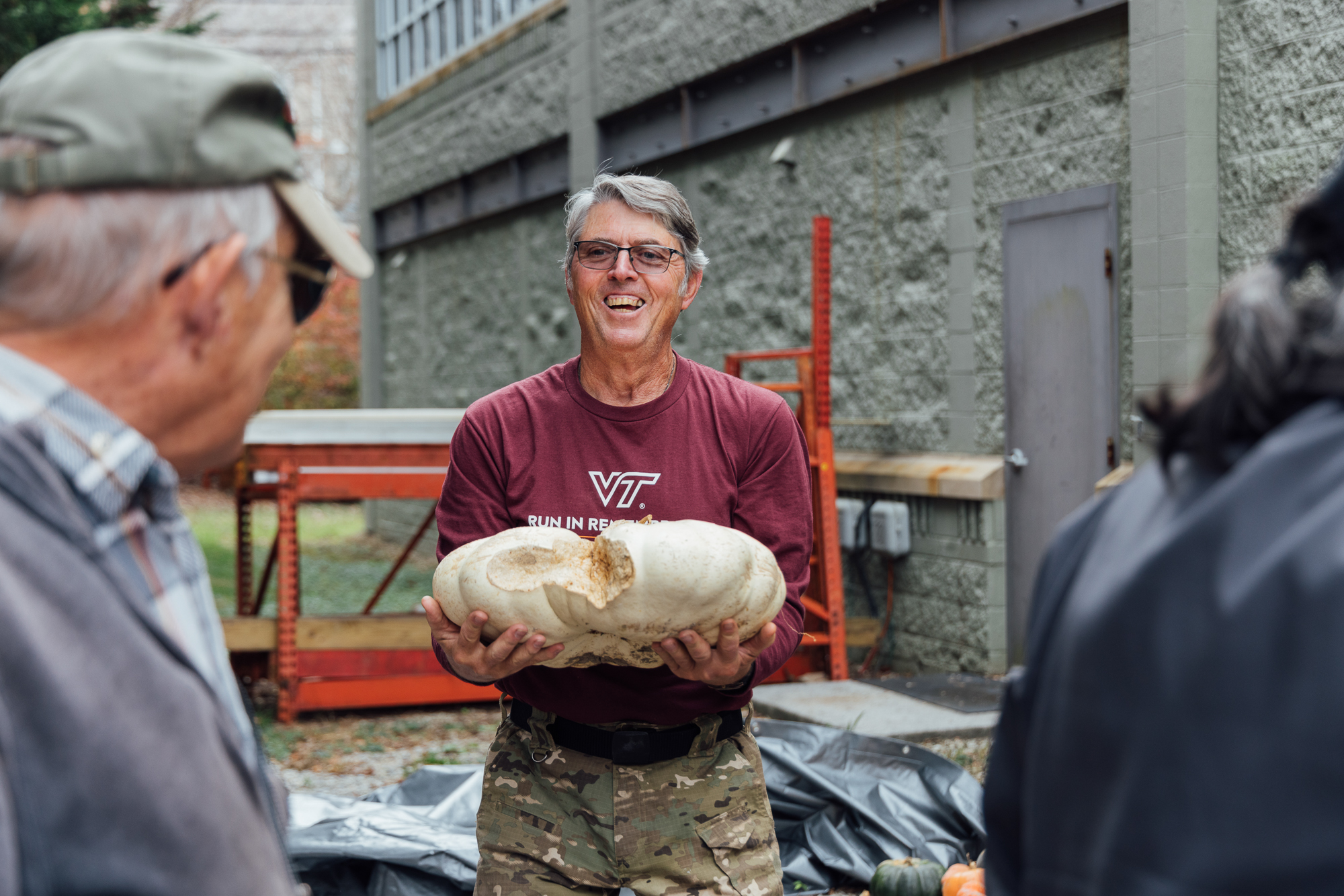 John Galbraith, wearing a maroon shirt and camouflage pants, holds a large white pumpkin, smiling while standing near a building with industrial features where the Glean Team stores some of its winter squash.