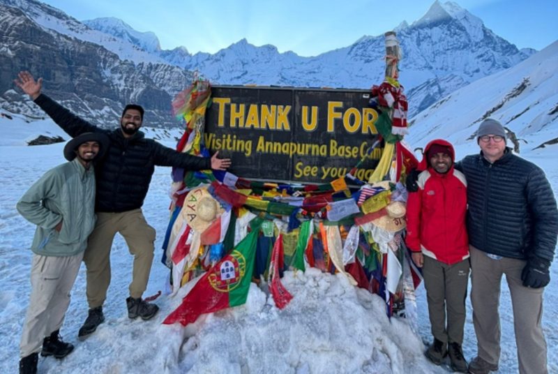 Four individuals gathered in the snowy, mountainous outdoors around a sign that says "Thank you for visiting the Annapura Base Camp."