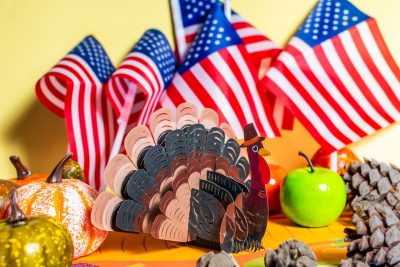 Mini American flags, pine cones, pumpkins, an apple, and a decorative paper turkey wearing a hat adorn a table.