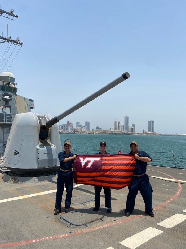 Three Navy officers stand on the forecastle of a military ship holding a striped VT flag. They are smiling and a large city sits across the water in the distance.