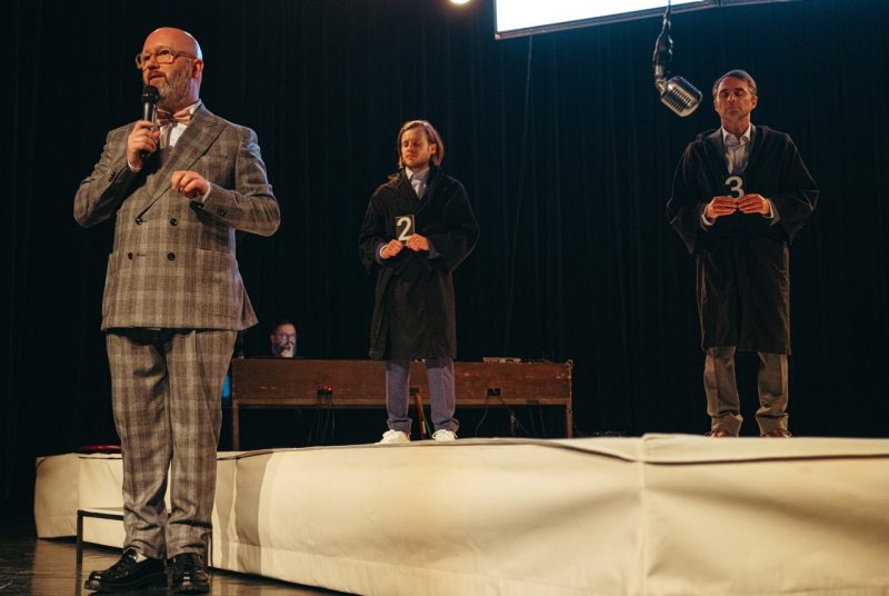 The cast of Ontroerend Goed's "Fight Night" performs on stage, two male candidates wearing black robes, one holding a sign with the number "2," the other holding number "3," are lined up on a platform while a white bald man in a plaid suit speaks into a microphone in front of them.