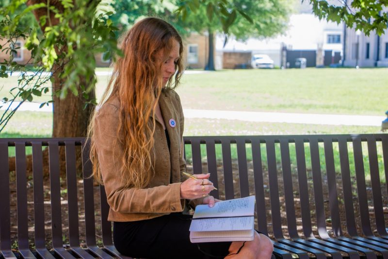 A young woman with long red hair sits on a bench under a tree and writes in her journal.