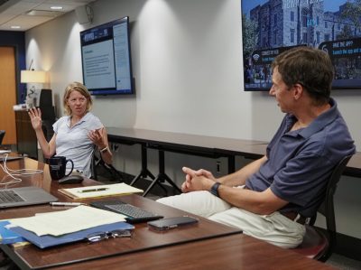 A woman and a man, both seated at a conference table surrounded by screens, are engaged in a discussion.