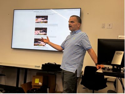 A man, wearing a blue and white striped shirt, stands in front of a screen as he presents.