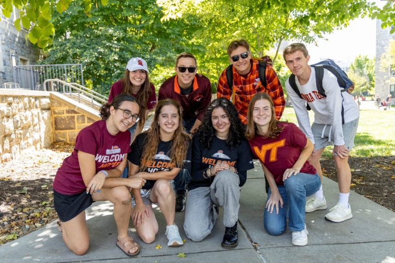 Residential Well-being Student Leaders smile and pose on the sidewalk outside of a Hokie Stone building.