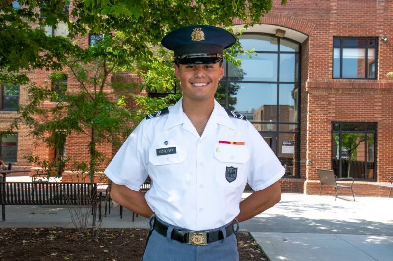 Cadet Schleiff stands in his uniform smiling in the shade of trees by a brick building.