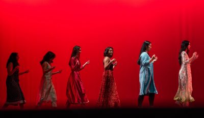 Women in dresses line up on stage silhouetted against a red background
