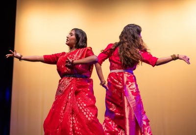 Two women dance on stage in colorful red saris 