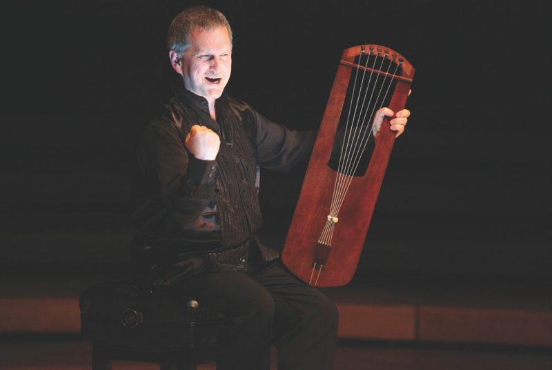 Medieval harpist Benjamin Bagby, a middle aged white man wearing a black shirt and pants, performs "Beowulf" while holding his harp in his lap. He emphasizes the line by making a fist with the other hand.
