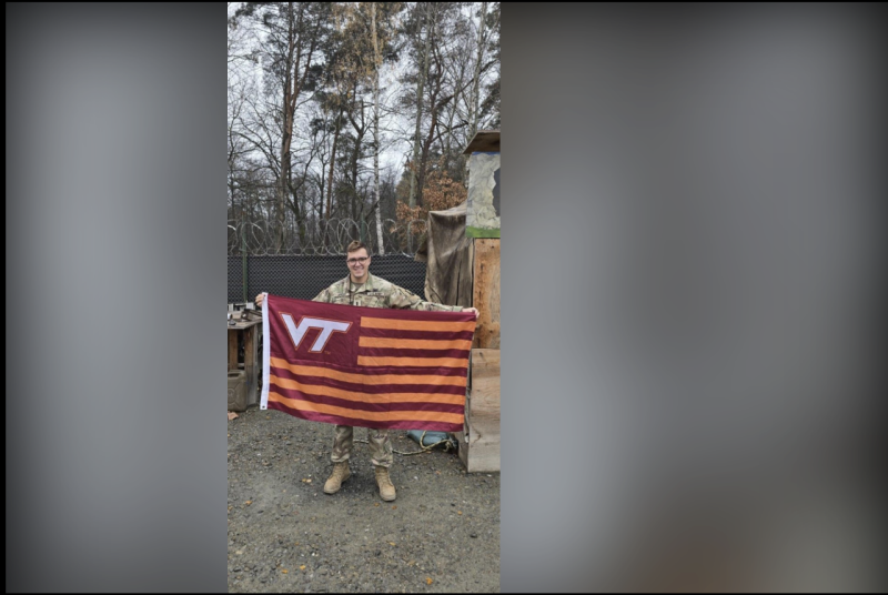 A serviceman in a camouflage uniform stands in front of barbed wire and chain link fencing holding a Virginia Tech flag. 