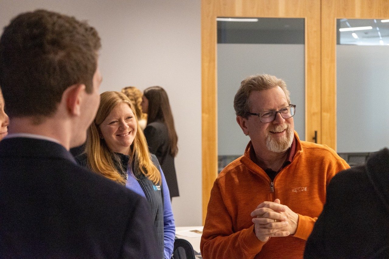 Man in glasses and an orange quarterzip wth a smiling woman in a vest over a purple long sleeve shirt.