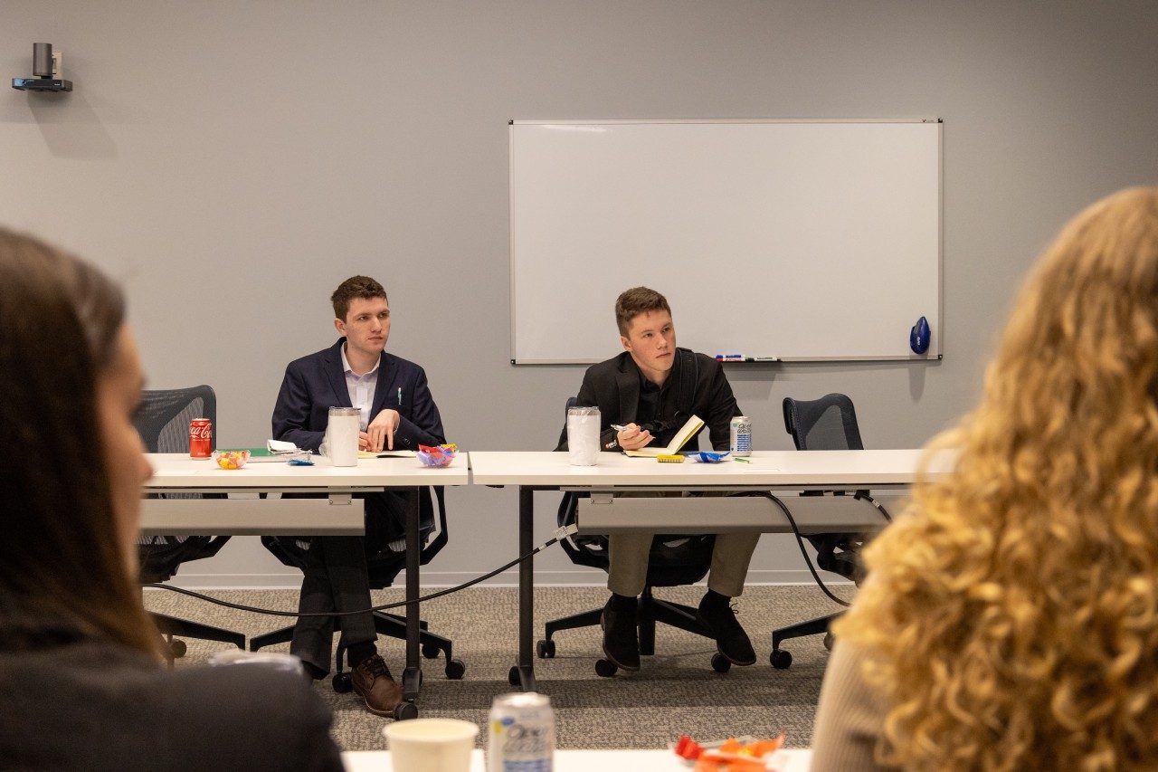 Two students sit at a table and listen to others speak.