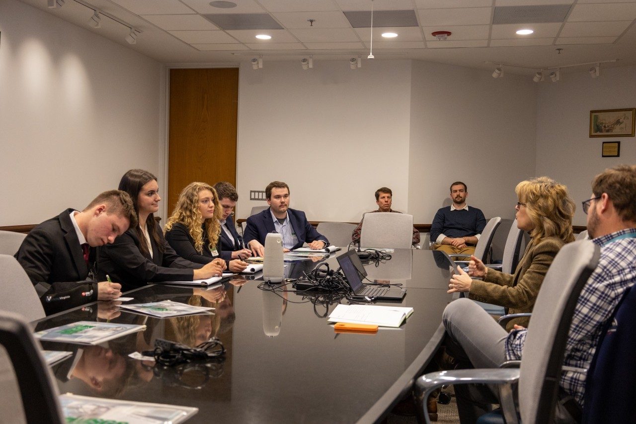 Students sit at a table and listen to a woman in a tan blazer speak.