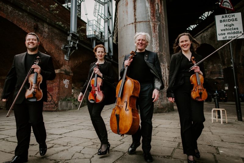 Members of the Dante Quartet hold their instruments in an industrial area of a city and smile widely.