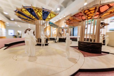 Within a wooden table-top model, small plastic figures of people stand on a circular surface, which is adjacent to another circular surface, both of which have circular roofs made of multi-colored and multi-patterned stained glass.