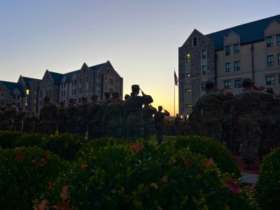 Cadets in uniform standing in groups salute as they are silhouetted by sunrise.