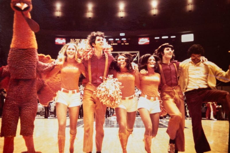 1973 long-necked HokieBird cheers with female and male cheerleaders on the basketball court. The female cheerleaders are wearing white hot pants, orange turtleneck with Hokies on it, and laced up orange knee high boots.