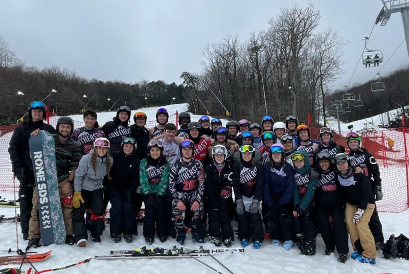 A group of 33 individuals dressed in skiing and snowboarding gear stand on a snowy mountain. Pairs of skis are in the snow in front of them, and a ski lift can be seen in the background.