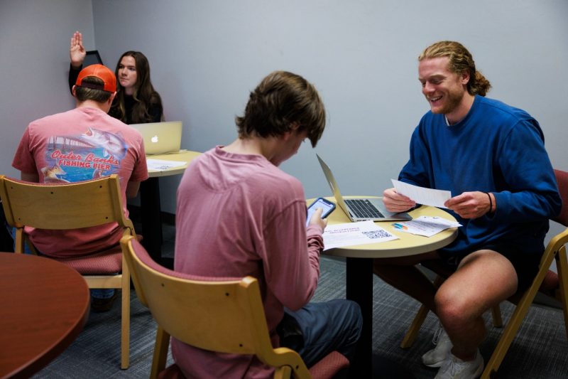 A young professional in a blue shirt sits across from a student in a maroon shirt at a small round table in a gray room. There is a laptop on the table, as well as a few scattered papers between them. The man in the blue sweater is smiling in mid conversation. The student is looking intently at something on his phone, held in his right hand. To their left, a young man in a maroon shirt and orange baseball hat and a young woman with long dark hair and a dark shirt sit at another small, round table. The woman's hand is raised as if she has a question.