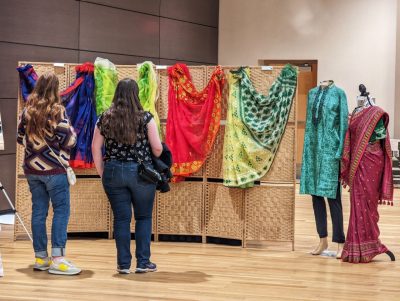 Two individuals stand facing a display backdrop on which four colorful garments are hung. The individuals are dressed in casual shirts, jeans, and sneakers, and each carries a small purse. To the right of the display, there are two mannequins dressed in colorful international attire: the one on the left is in a turquoise dress with dark pants, and the one on the right wears a maroon, orange, and green dress with a maroon and orange covering.
