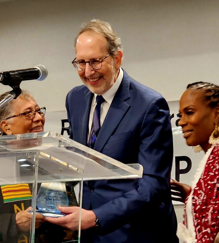 A man in a suit stands at a podium and is handed an award from a woman.