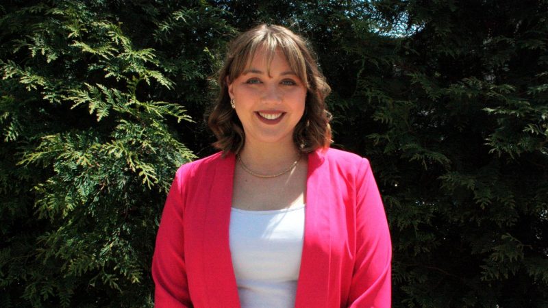 A woman in a pink jacket stands in front of foliage.