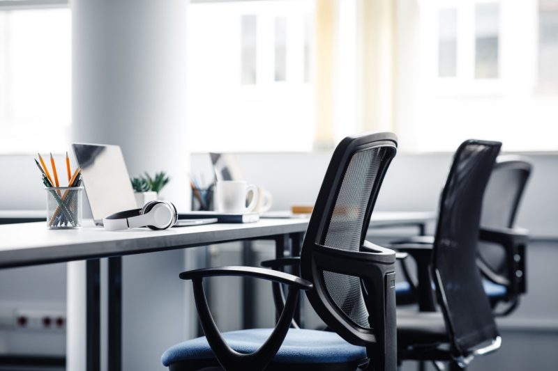 Three desks and desk chairs side by side in an office. Atop them are computers, a pair headphones, three cups of coffee, and containers of pens. In the background is a window.