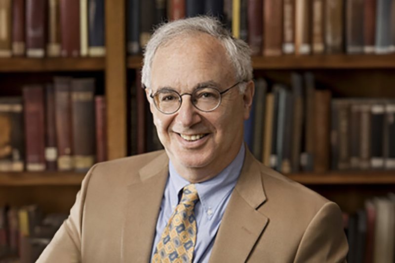 A man is sitting in front of a bookshelf. He is wearing glasses and a tan suit with a blue shirt and yellow and blue tie.