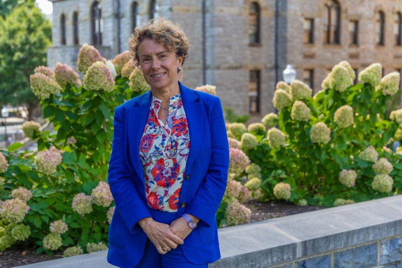 A woman wearing a bright blue suit and a floral blouse stands with her hands clasped in front of pink  and yellow hydrangeas. 