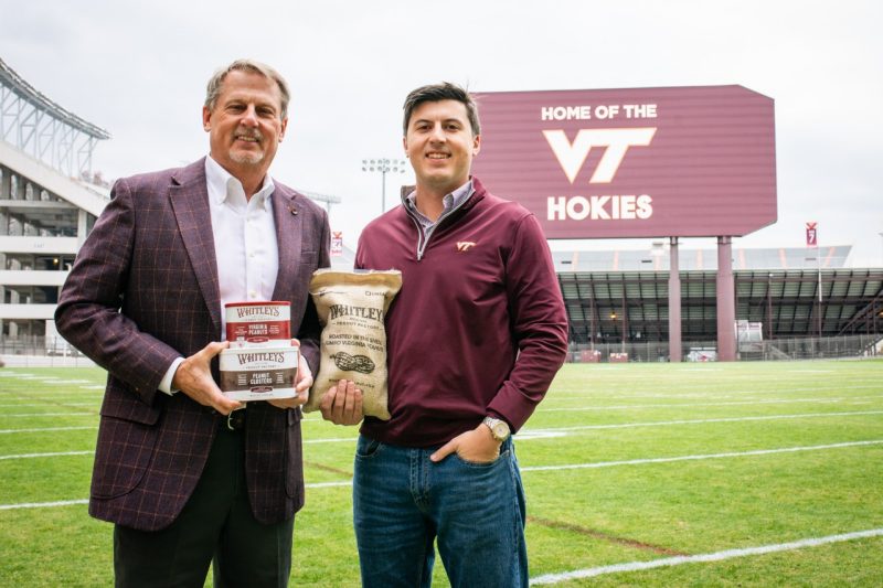 Two men stand on a football field in front of a sign holding food products.