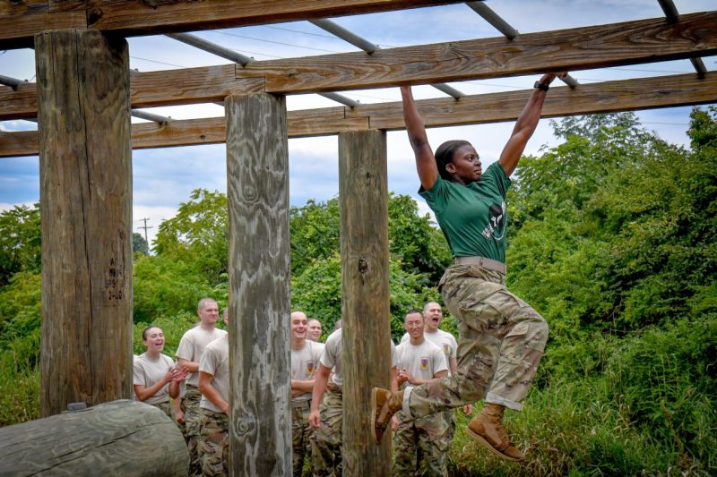 A woman cadet in a military uniform swings from a monkey bar obstacle and looks determined as she crosses it successfully. Cadets cheer in the background.