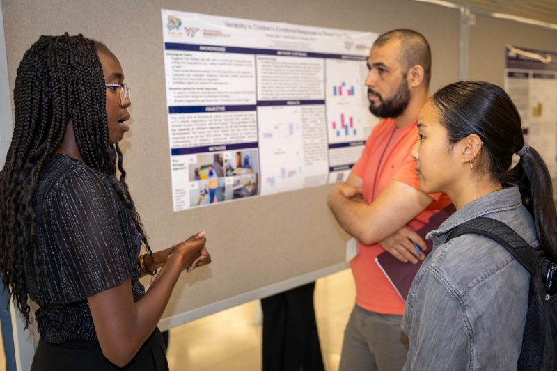 A student stands to the left discussing a research poster with two individuals. 