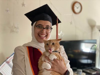 A young woman in graduation attire holding an orange American Shorthaired cat. The cat is licking her finger.