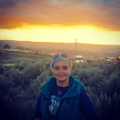 Person with blue hair standing outside with a sunset in the background.
