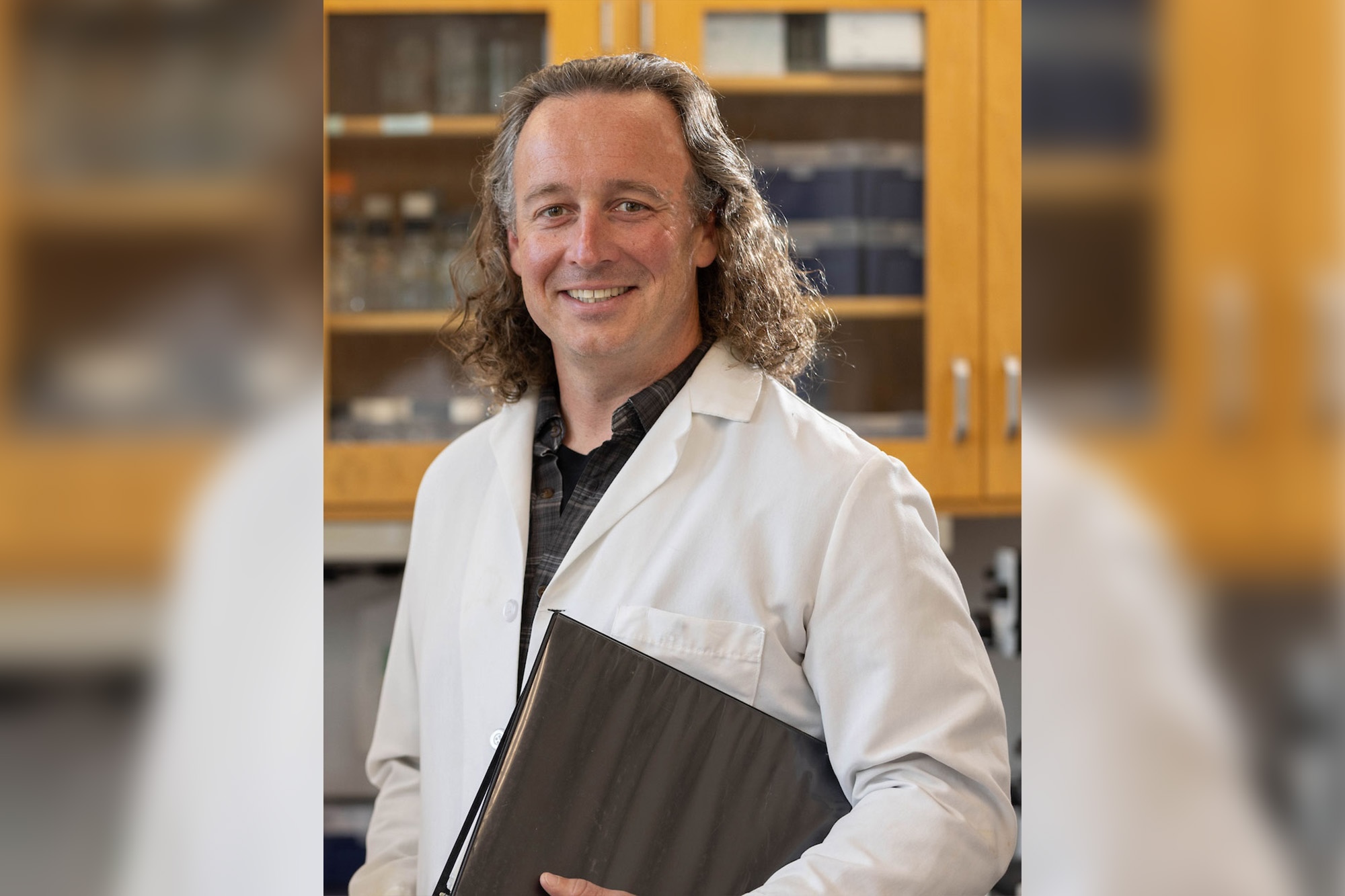 Clayton Caswell posing for a headshot wearing a white lab coat and holding a black binder in his laboratory