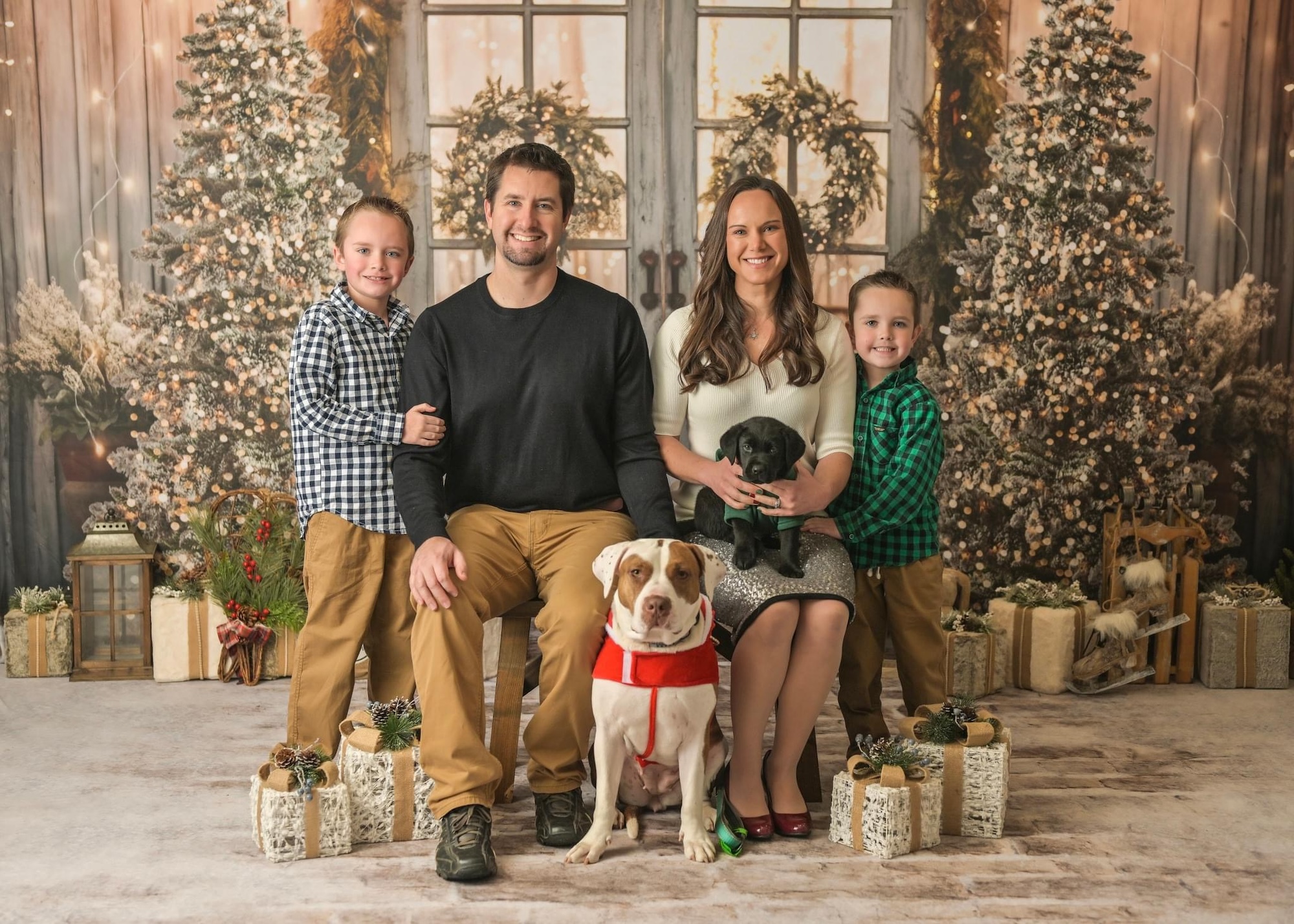 Virginia Edwards (at right) posing for a family photo with her husband (at left), two sons (far right and far left), and two dogs (at front). The family is posing in front of two Christmas trees with prop presents under them.