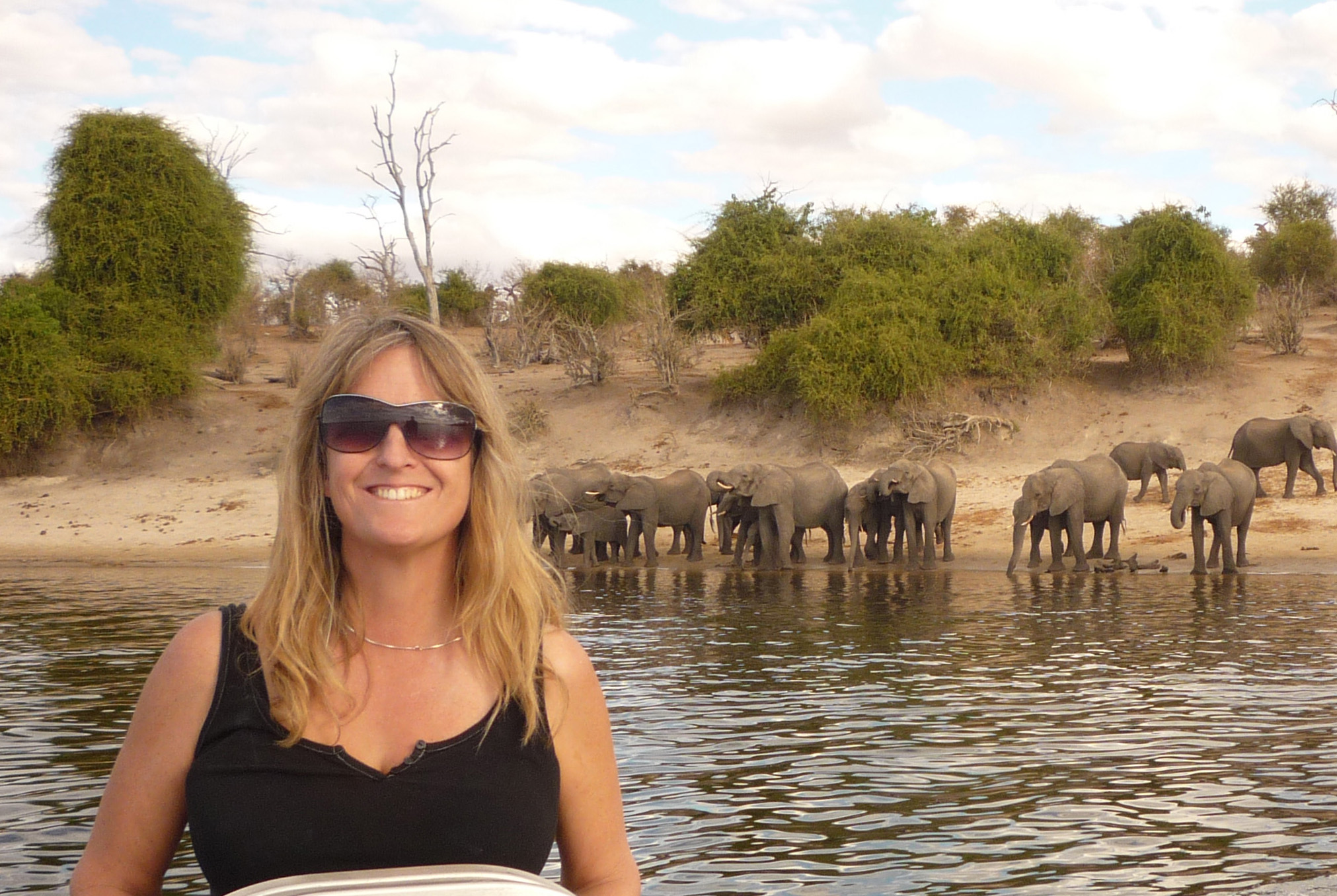 Kathleen Alexander at the helm of a small boat with elephants standing on the shore in the background