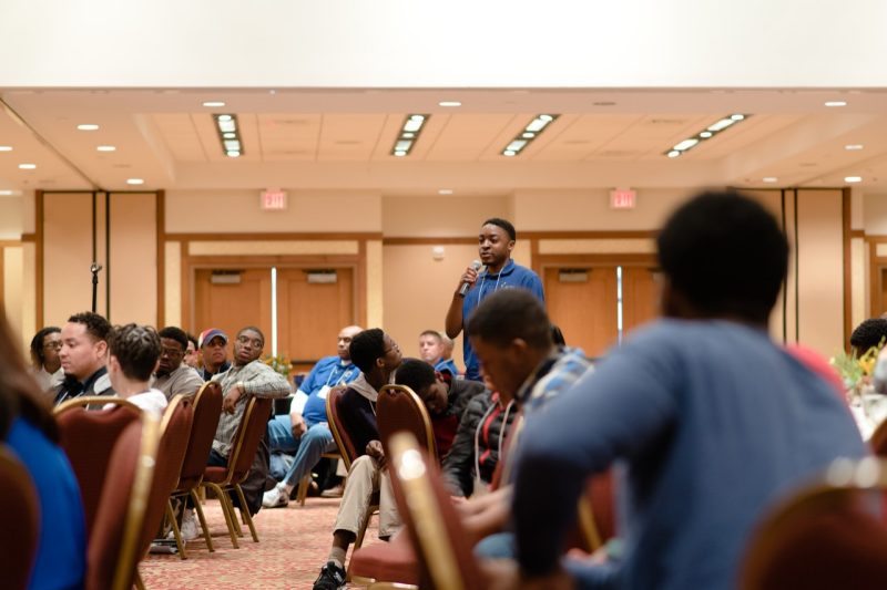 A Black man holding a microphone while participants in the 2018 Uplifting Black Men Conference look on.