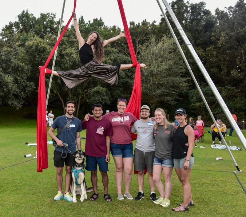 Six people pose below an aerialist high in the air between two red silk ropes.