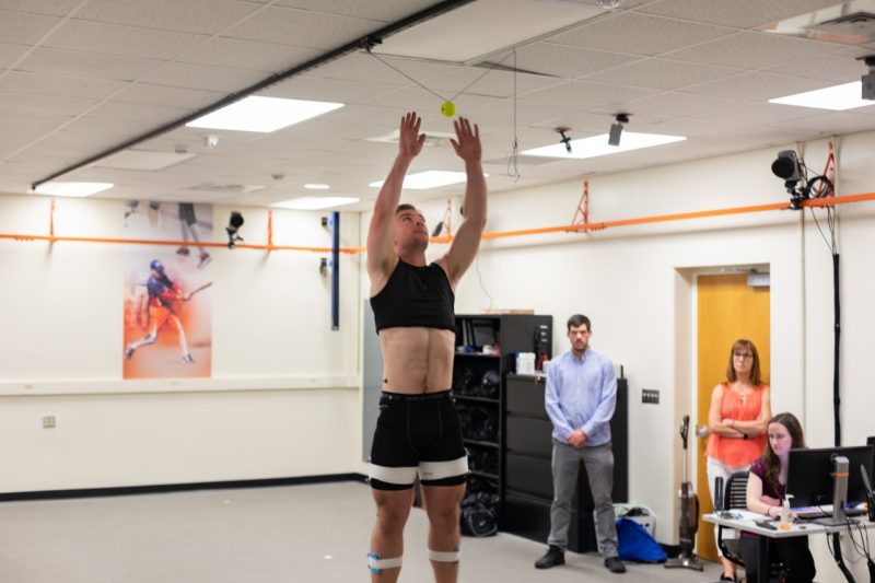 Male participant in cropped black shirt and athletic shorts jumps, with his hands in the air, and researchers observe in the background.