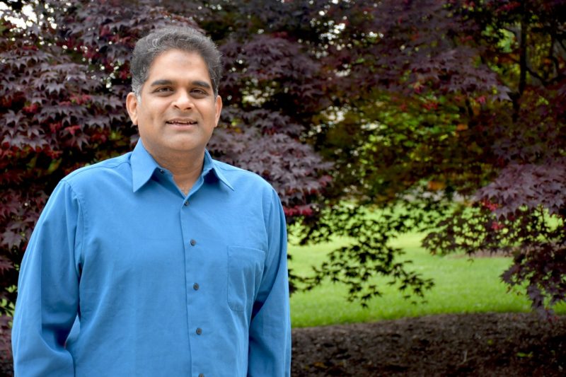 A man with dark hair and wearing a button down blue dress shirt stands in front of a bush with red leaves.