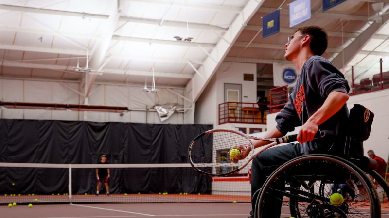 A player in a wheelchair prepares to serve the ball to member of the Virginia Tech men’s tennis team standing on the other side of the net.