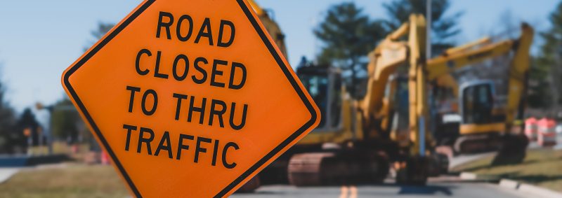 An orange road closed sign leans into view, blocking a campus road for construction activity in the background.